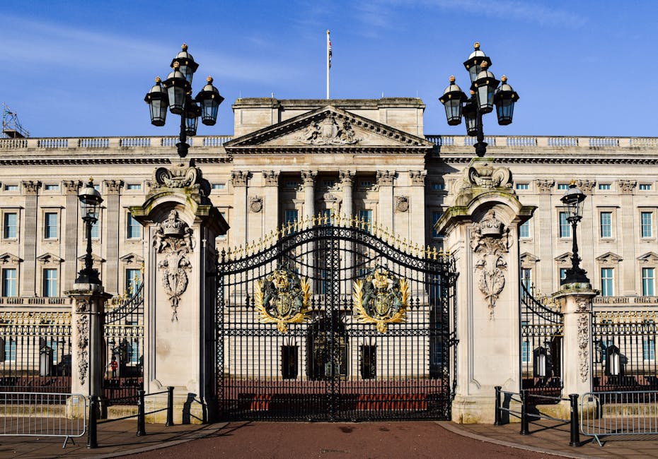 A large, ornate wrought iron gate with decorative gold accents and intricate patterns, set on tall white stone pillars topped with elaborately designed lanterns, providing access to the grounds of Buc