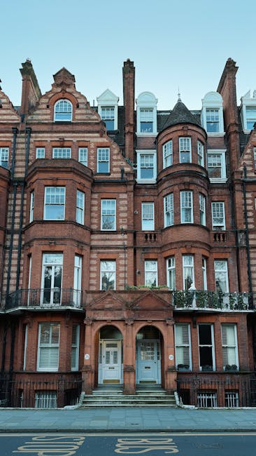 A tall, multi-storey red-brick building with large white-framed windows and decorative architectural details, situated on a street with a sidewalk lined by blossoming trees. The building features orna